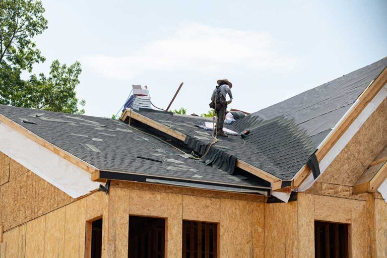 Two people standing on ladders cleaning debris from a gutter along a shingled roof in early spring.