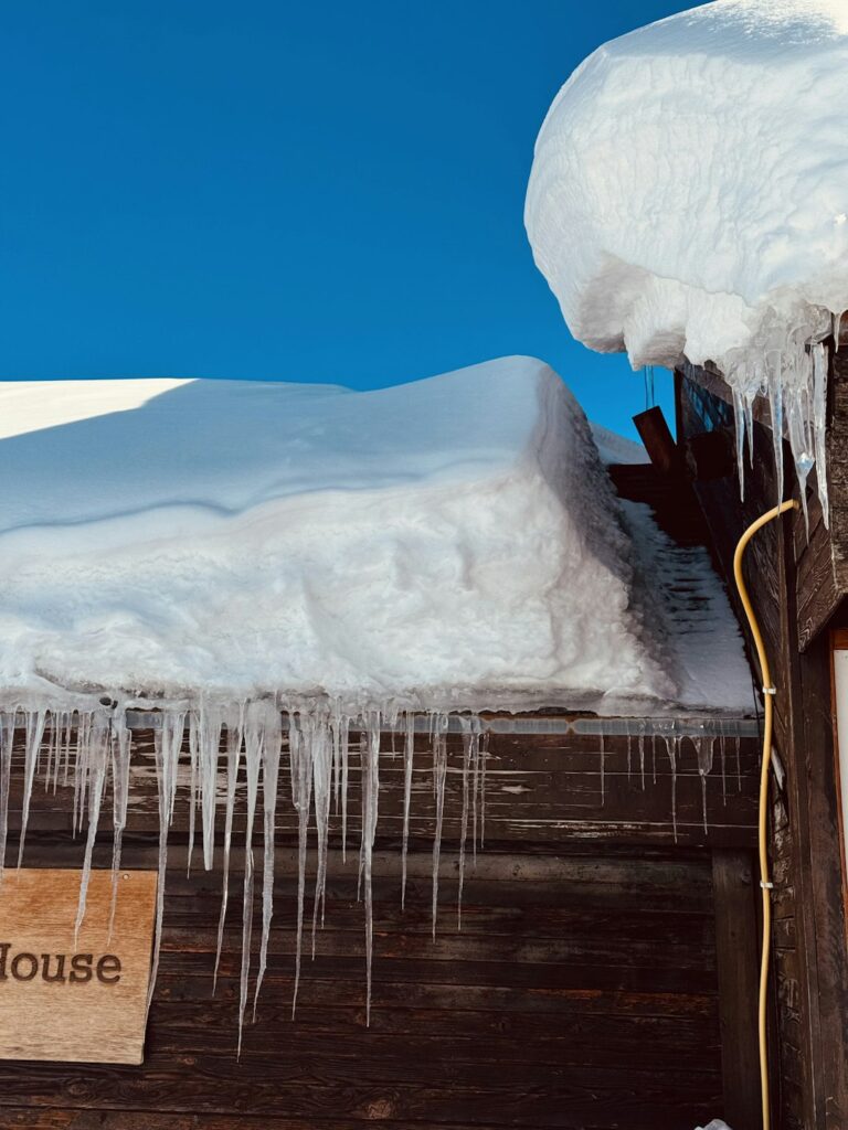Snow-covered residential roof with an ice dam, hanging icicles, and water staining visible near the attic edge.