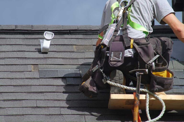 A homeowner inspects a rain-filled gutter beneath a shingled roof with a downspout beside the wall.